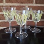 Fire klare aperitifglas med grønt rombeformet mønster, stående på en mørk overflade foran en murstensvæg.