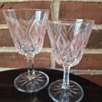 Et par klare VMC Reims aperitifglas med et præget mønster i glasset, stående på en mørk overflade foran en murstensvæg.