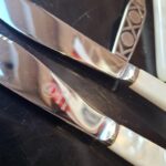 Et arrangement af vintage gafler og smørknive med perlemorsgreb og mønstrede stålhåndtag, sammen med fire blå-hvide porcelænsholdere.