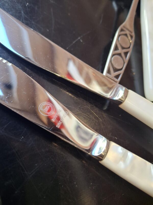 Et arrangement af vintage gafler og smørknive med perlemorsgreb og mønstrede stålhåndtag, sammen med fire blå-hvide porcelænsholdere.