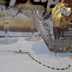 Et sæt vintage krystalglas og en krystalskål med chokolade, elegant arrangeret på en hvid dug med broderede blomster.