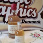 Nærbillede af Chocolate Graphics Danmark skiltet bag to flasker Julemjød i forskellige størrelser, med krystalglas i forgrunden.
