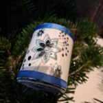 Et lille, håndlavet, rødt juletrummeophæng dekoreret med et hvidt/sølvbånd med kristtjørneblade og guldsnor. Hænger i juletræets grangrene.
