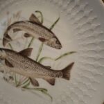 Nærbillede af en hvid Bavaria Schumann Arzberg tallerken med reliefkant og et motiv af to hvidfisk omgivet af vandplanter.