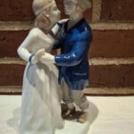 Et porcelænsfigur af et elegant dansende par, hvor manden bærer en grå hat og en mørkeblå frakke, og kvinden en hvid kjole.