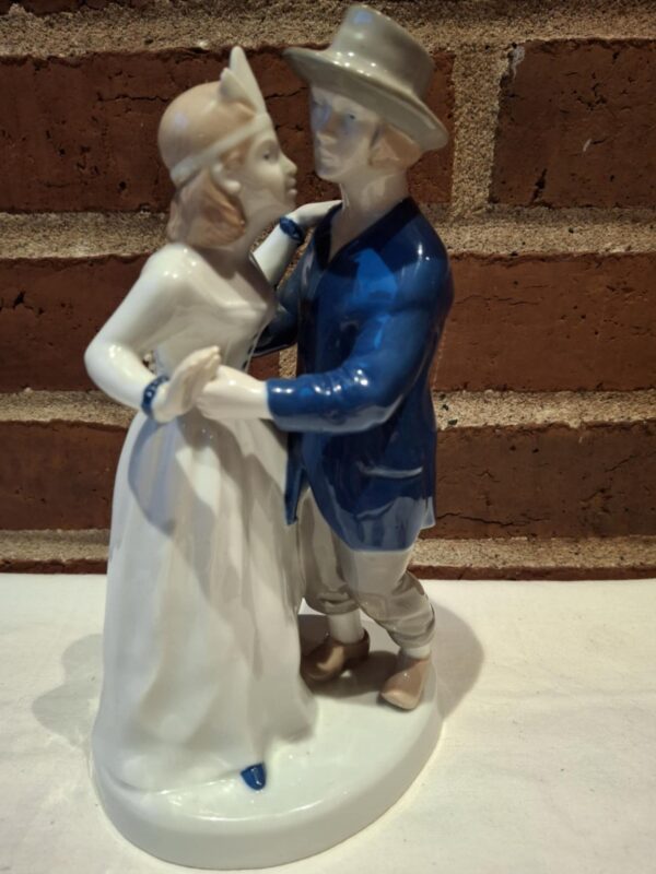 Et porcelænsfigur af et elegant dansende par, hvor manden bærer en grå hat og en mørkeblå frakke, og kvinden en hvid kjole.