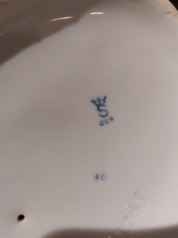 Billede af bunden af en porcelænsfigur, der viser mærket en krone med et 'S' og teksten 'GOR' nedenunder i blåt.