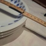 Et målebånd der måler bredden på en Lyngby Danild 'Dråben' tallerken i hvid porcelæn med gråt og blåt dråbemønster.