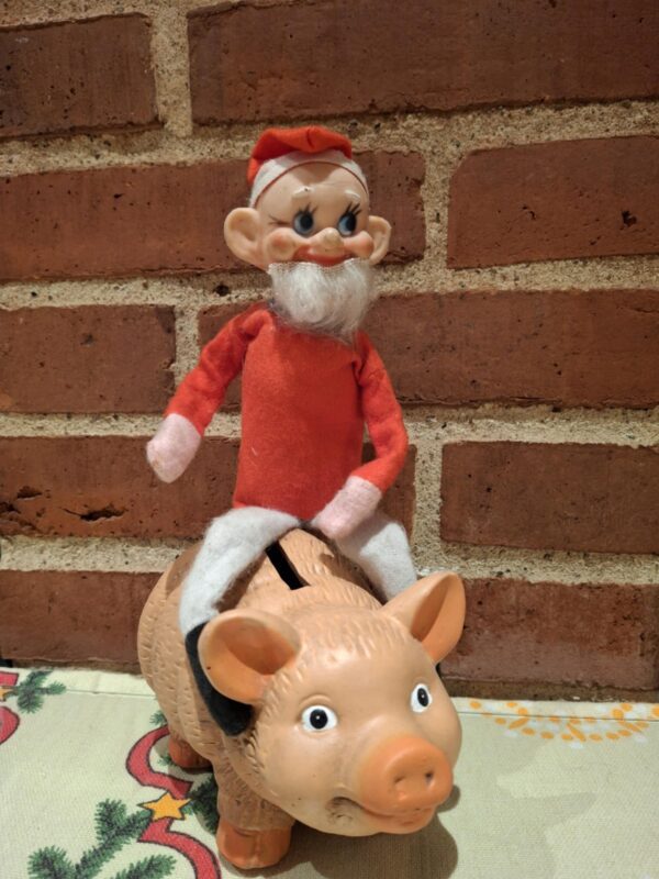En retro julenisse figur med rød beklædning og hvidt skæg, siddende på en terracotta sparegris.