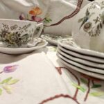 En hvid porcelænskaffekop og underkop med et detaljeret blomster- og fuglemønster i brune og grønne nuancer, stående på en broderet dug.