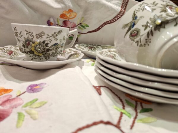 En hvid porcelænskaffekop og underkop med et detaljeret blomster- og fuglemønster i brune og grønne nuancer, stående på en broderet dug.