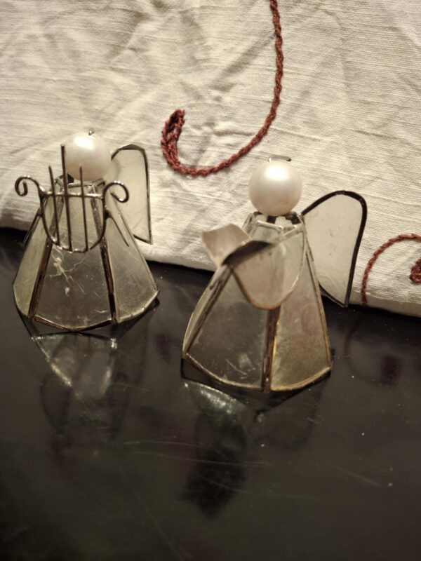 To små englefigurer lavet af glas og metal, hvor den ene holder en harpe, stående på et mørkt, reflekterende underlag.