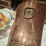 Nærbillede af det patina-fyldte kobberhåndtag på en vintage champagnekøler, der fremhæver messingdetaljer og nitter. En champagneflaske kan anes i den slørede baggrund.