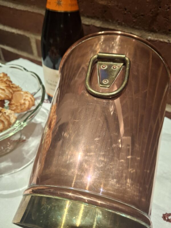 Nærbillede af det patina-fyldte kobberhåndtag på en vintage champagnekøler, der fremhæver messingdetaljer og nitter. En champagneflaske kan anes i den slørede baggrund.