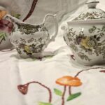Et engelsk porcelænssæt bestående af flødekande og sukkerskål i hvid med et mønster af blomster og fasaner, stående på en hvid dug.