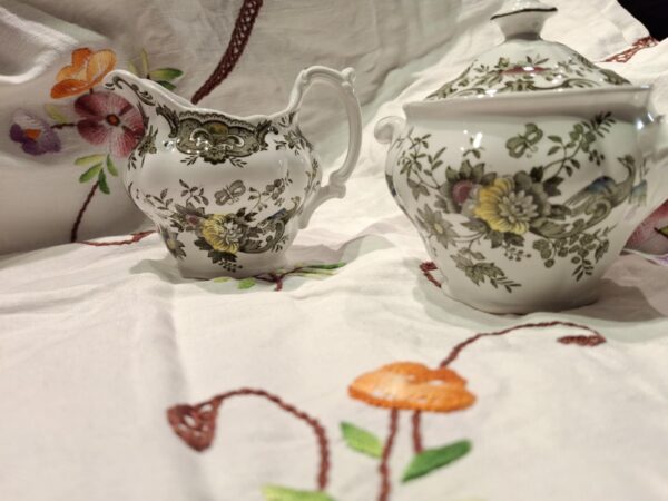 Et engelsk porcelænssæt bestående af flødekande og sukkerskål i hvid med et mønster af blomster og fasaner, stående på en hvid dug.