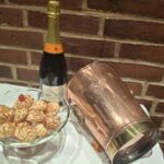 En skinnende vintage champagnekøler i kobber og messing (22 cm høj), placeret skråt ved siden af en champagneflaske og en glasskål med småkager på en hvid dug foran en murstensvæg.