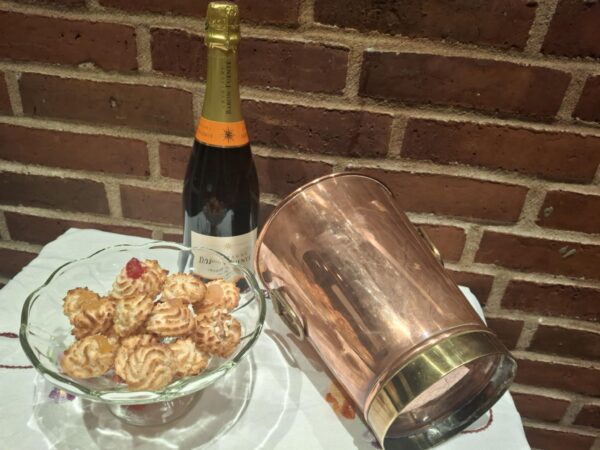 En skinnende vintage champagnekøler i kobber og messing (22 cm høj), placeret skråt ved siden af en champagneflaske og en glasskål med småkager på en hvid dug foran en murstensvæg.
