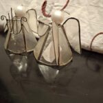 Nærbillede af to små englefigurer af gennemsigtigt glas og metal, set forfra og let skråt, på et mørkt bord.