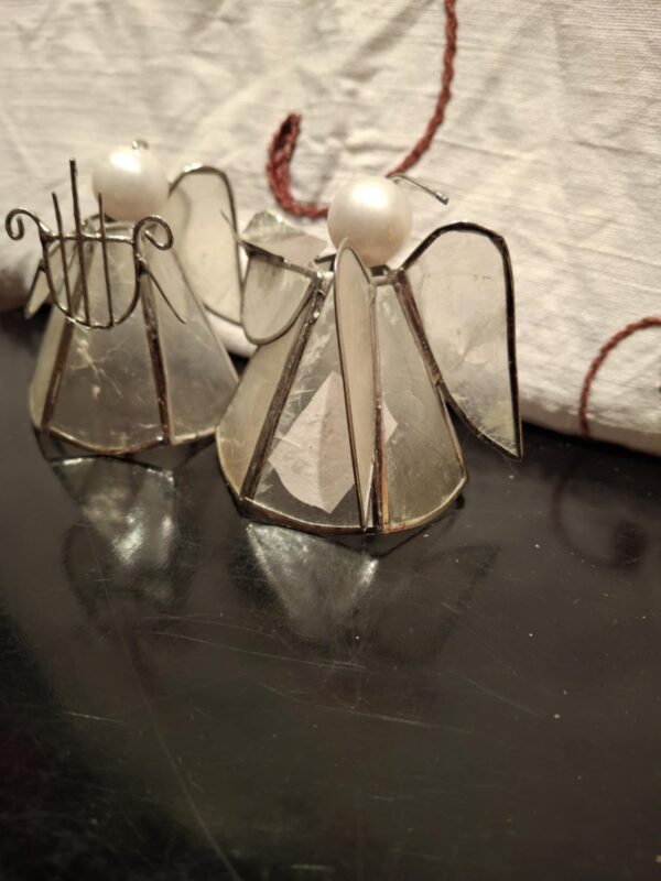 Nærbillede af to små englefigurer af gennemsigtigt glas og metal, set forfra og let skråt, på et mørkt bord.