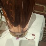 Nærbillede af bunden af en vintage kobber champagnekøler (22 cm høj), der viser rustikke detaljer som nitter og sammenføjninger, placeret på en hvid dug med broderede blomster.