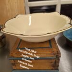 Oval cremefarvet Johnson Bros. 'Virton' skål med elegant guld- og sort kant, placeret på en rustik træboks. Teksten '1 fad /skål kartoffelskål, frugt. nøgler mm. virtorian johnson bros England' er synlig.