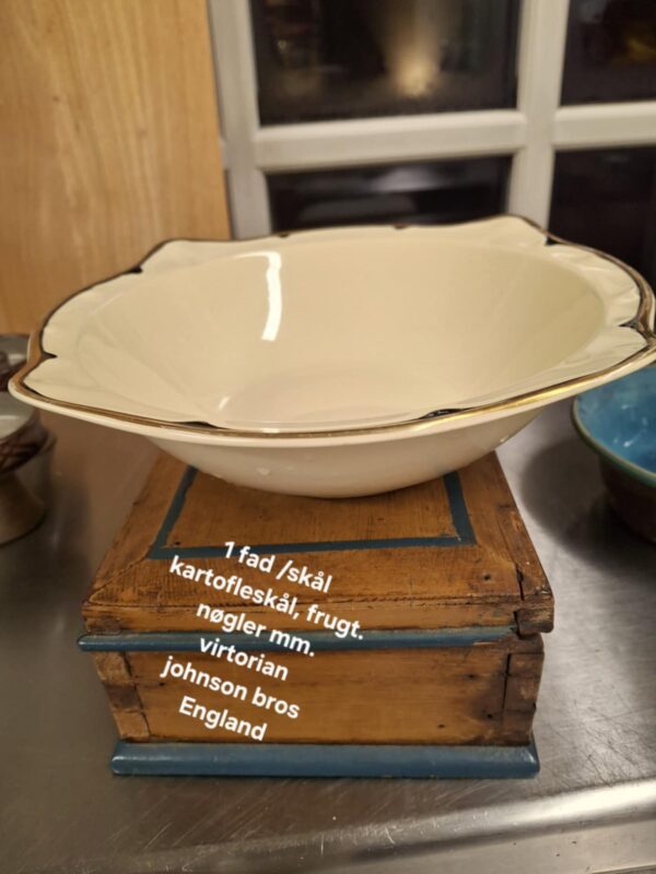 Oval cremefarvet Johnson Bros. 'Virton' skål med elegant guld- og sort kant, placeret på en rustik træboks. Teksten '1 fad /skål kartoffelskål, frugt. nøgler mm. virtorian johnson bros England' er synlig.
