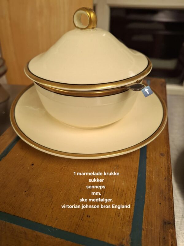 Sart creme marmeladekrukke med låg og tilhørende underskål fra Victorian Johnson Bros., dekoreret med guldkant. Set fra siden med hvid tekst: '1 marmelade krukke sukker senneps mm. ske medfølger. virtorian johnson bros England'.