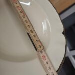 Nærbillede af en dyb tallerken fra Johnson Bros. 'Virtorian Cream' med guldkant, der viser en tommestok, der indikerer en diameter på ca. 19 cm, og fanger detaljerne i den bølgede kant.