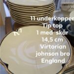 En enkelt cremehvid Johnson Bros. 'Victorian' underkop med guldkant, der er placeret foran en stabel lignende underkopper. Der er tekst på billedet, der angiver antal, mål og mærke, samt info om et skår.