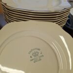 Bagsiden af en cremefarvet, antikke tallerken fra Johnson Bros. med tydeligt grønt stempel 'Victorian Johnson Bros Made in England', omgivet af en kongekrone. En stabel lignende tallerkener anes i baggrunden.