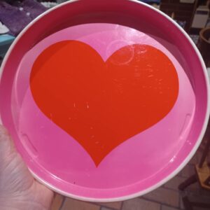 Et rundt, pink serveringsfad med et stort rødt hjerte i midten, holdt i hånden af en person, mod en murstensvægindendørs.
