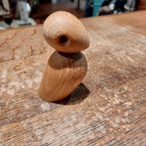 En lille, stiliseret træfugl med et aflangt hoved og krop, udskåret i lyst træ med synlige åretegninger, stående på et bord.