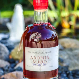 En flaske Ny Nordisk Mjød Aronia Mjød 35 CL med rød voksforsegling, placeret udendørs på en sten i sollys.