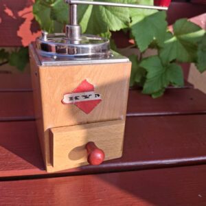 En gammel kaffekværn i lyst træ med metaltop og et 'KYN'-logo. Den har en rød knop på håndtaget og på skuffen, udendørs.