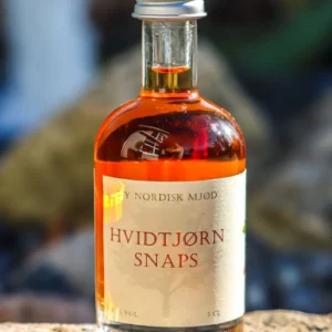 En flaske 'NY NORDISK MJØD HVIDTJØRN SNAPS' på 5 cl med ravfarvet væske, placeret udendørs på en sten med sløret grøn baggrund.