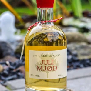 En flaske 'NY NORDISK MJØD JULE MJØD' på 35 cl med lys gylden væske og rød forsegling, placeret udendørs på en sten ved en vandfontæne.