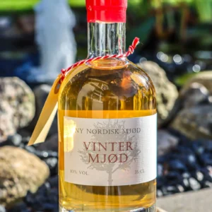 En flaske 'NY NORDISK MJØD VINTER MJØD' på 35 cl med lys gylden væske og rød forsegling, placeret udendørs på en sten ved en vandfontæne.
