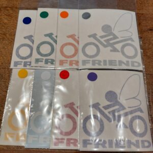 Et guld Bikerfriend Skyts Fe klistermærke i gennemsigtig plastikpakke med en gul cirkel på label, placeret på et træbord.