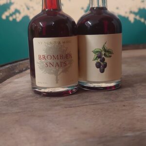 To små flasker 'NY NORDISK MJØD BROMBÆR SNAPS' på 5 cl med mørk væske, stående på en træoverflade.