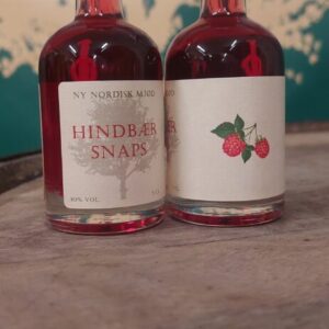 To små flasker 'NY NORDISK MJØD HINDBÆR SNAPS' på 5 cl med rødlig væske, stående på en træoverflade.