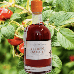 En flaske 'NY NORDISK MJØD HYBEN MJØD' på 35 cl med rødlig væske og orange voksforsegling. I baggrunden ses grønne hybenbuske.
