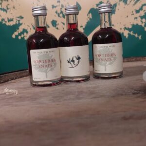 Tre små flasker 'NY NORDISK MJØD SORTEBÆR SNAPS' på 5 cl med mørk væske, stående på en træoverflade med grønt baggrundstæppe.