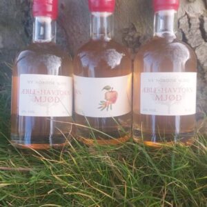 Tre flasker 'NY NORDISK MJØD ÆBLE-HAVTORN MJØD' på 35 cl med lyserød væske og rød vokslignende forsegling, stående på græs ved et træ.