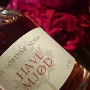 Nærbillede af en flaske 'Ny Nordisk Mjød HAVE MJØD' på 35 cl med en rødlig væske, foran røde blomster.
