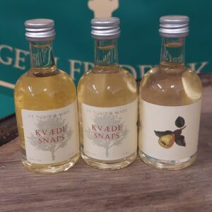 Tre små flasker 'NY NORDISK MJØD KVÆDE SNAPS' på 5 cl med gullig væske, stående på en træoverflade med grønt baggrundstæppe.