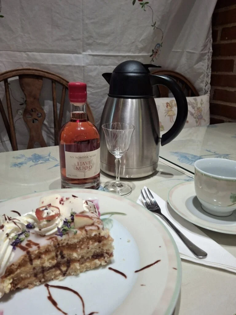 Et stykke lagkage, en kaffekop og en flaske Have Mjød på et dækket bord