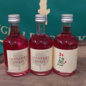 Tre små flasker 'NY NORDISK MJØD TRANEBÆR SNAPS' på 5 cl med rødlig væske, stående på en træoverflade med grønt baggrundstæppe.