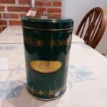 En høj, grøn retro kaffedåse med gulddetaljer og ordet 'Coffee' på en gylden plakette, stående på et blomstret bord.