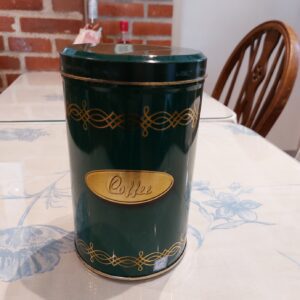 En høj, grøn retro kaffedåse med gulddetaljer og ordet 'Coffee' på en gylden plakette, stående på et blomstret bord.